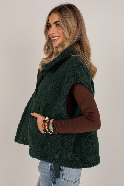 A woman wearing a dark forest green sherpa fleece vest over a brown long-sleeve top and light blue jeans, posing against a neutral light gray background.