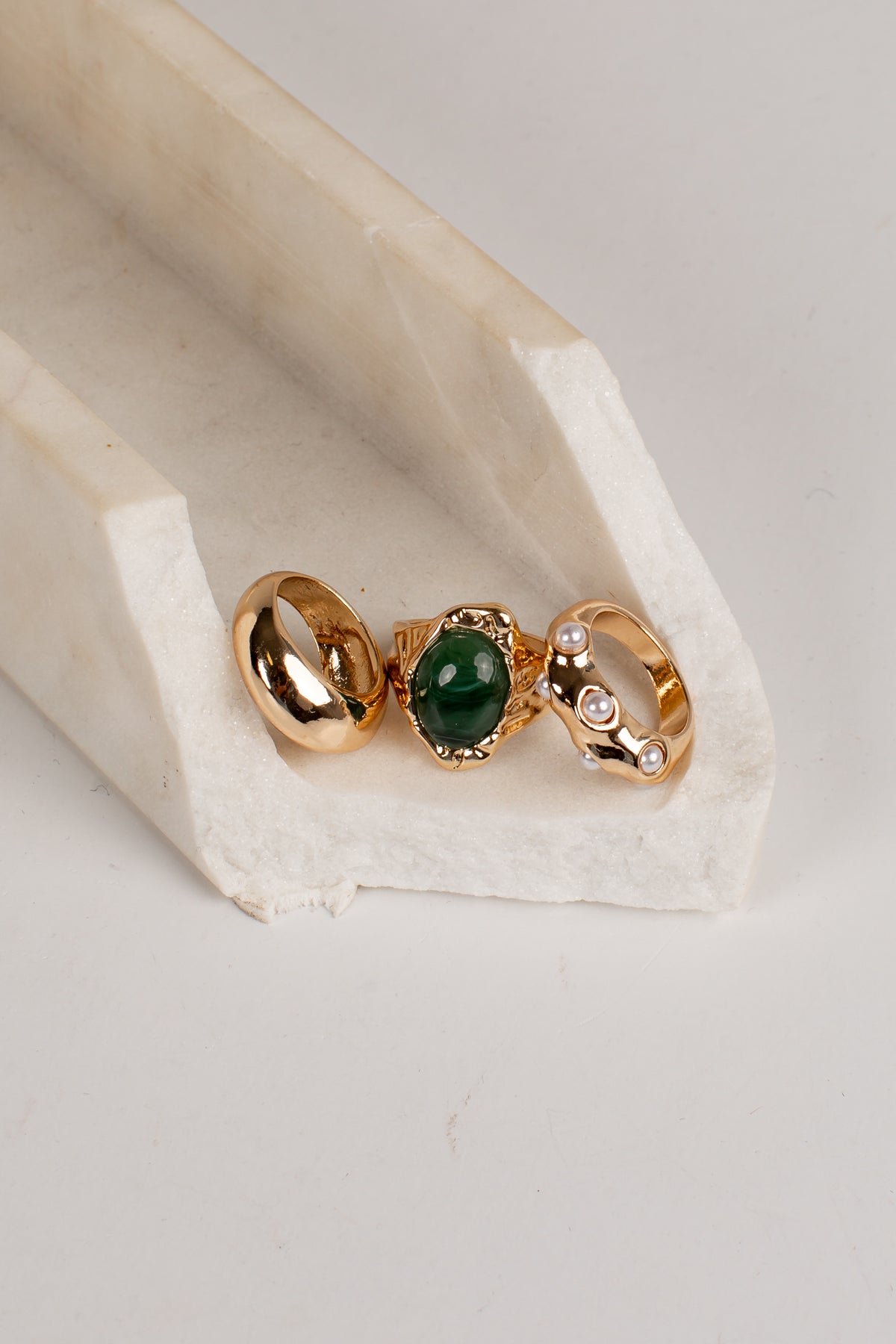 Three gold rings displayed on a white stone stand against a light gray background. The left ring is smooth gold, the center ring features a green oval stone in a gold setting, and the right ring has a gold band with small clear stones.