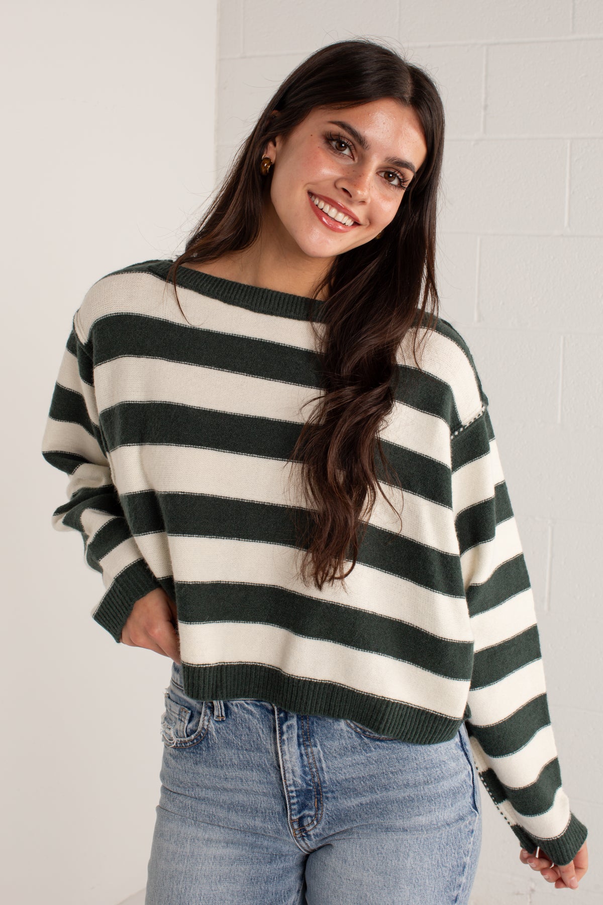 A woman wearing a dark green and cream horizontal striped knit sweater with light wash blue jeans, standing against a white brick wall.