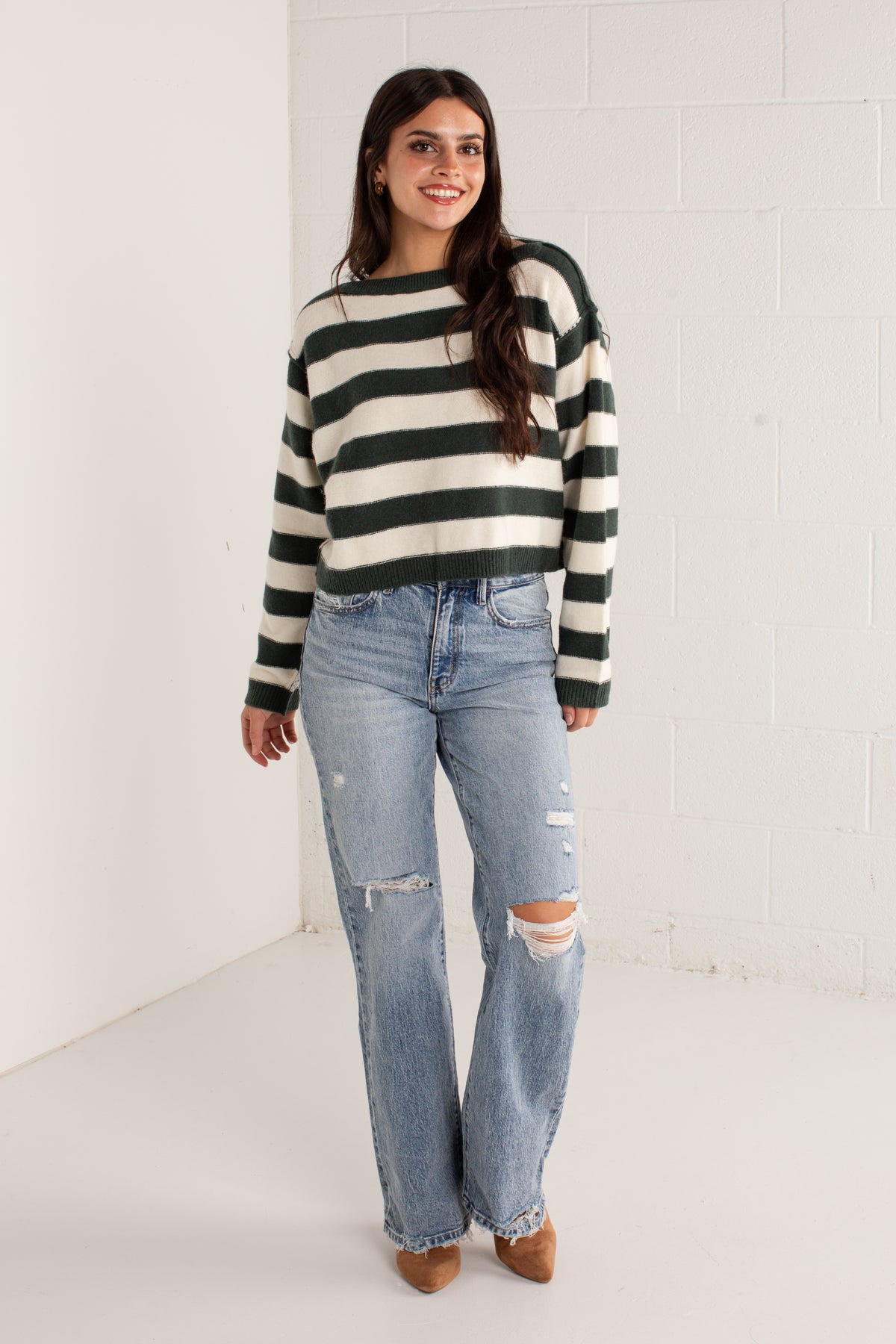 A woman modeling a dark green and cream striped knit sweater paired with light-wash distressed jeans and brown pointed-toe heels, standing against a white brick wall.