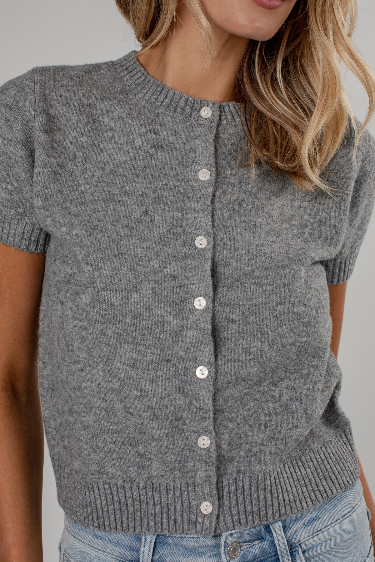A close-up of a woman wearing a short-sleeve gray knit cardigan with white buttons, paired with light blue jeans, against a plain light gray background.