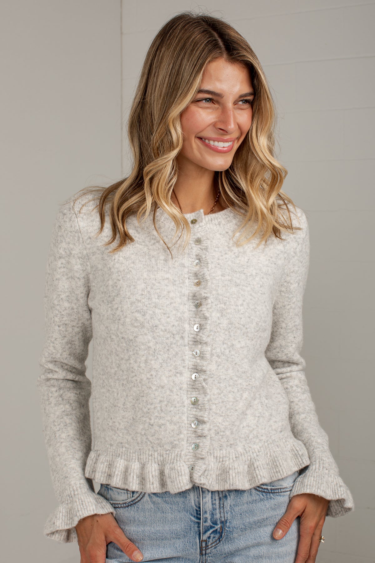 A woman wearing a light heather gray knit cardigan with a button-down front and ruffled hem, paired with blue jeans, against a neutral gray background.