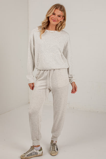 A woman modeling a light heather gray two-piece loungewear set consisting of a long-sleeve sweatshirt and matching jogger pants, paired with distressed sneakers, in a bright studio setting.
