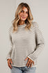 A woman wearing a beige and gray horizontally striped knit sweater with light blue jeans, standing against a light gray brick wall.