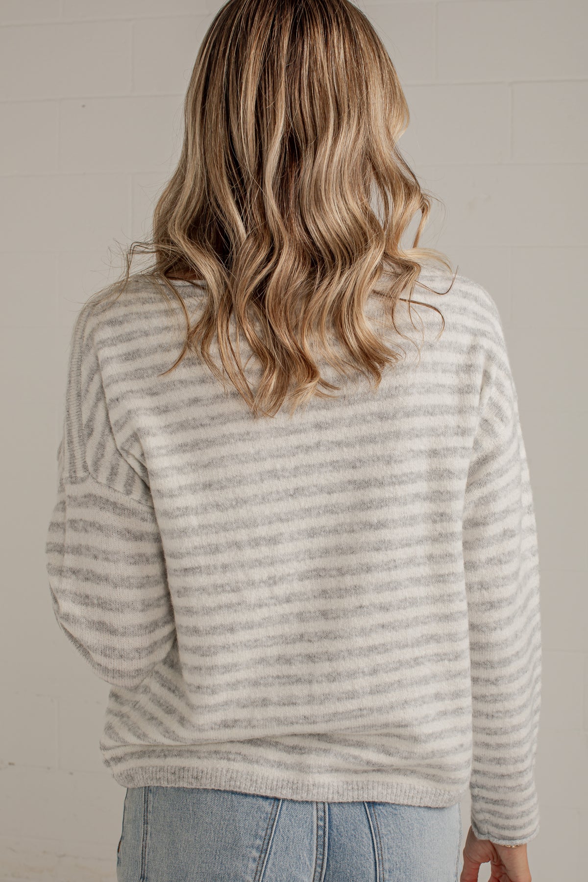 A woman viewed from behind wearing a light gray and white horizontally striped knit sweater and light blue jeans against a neutral off-white wall.