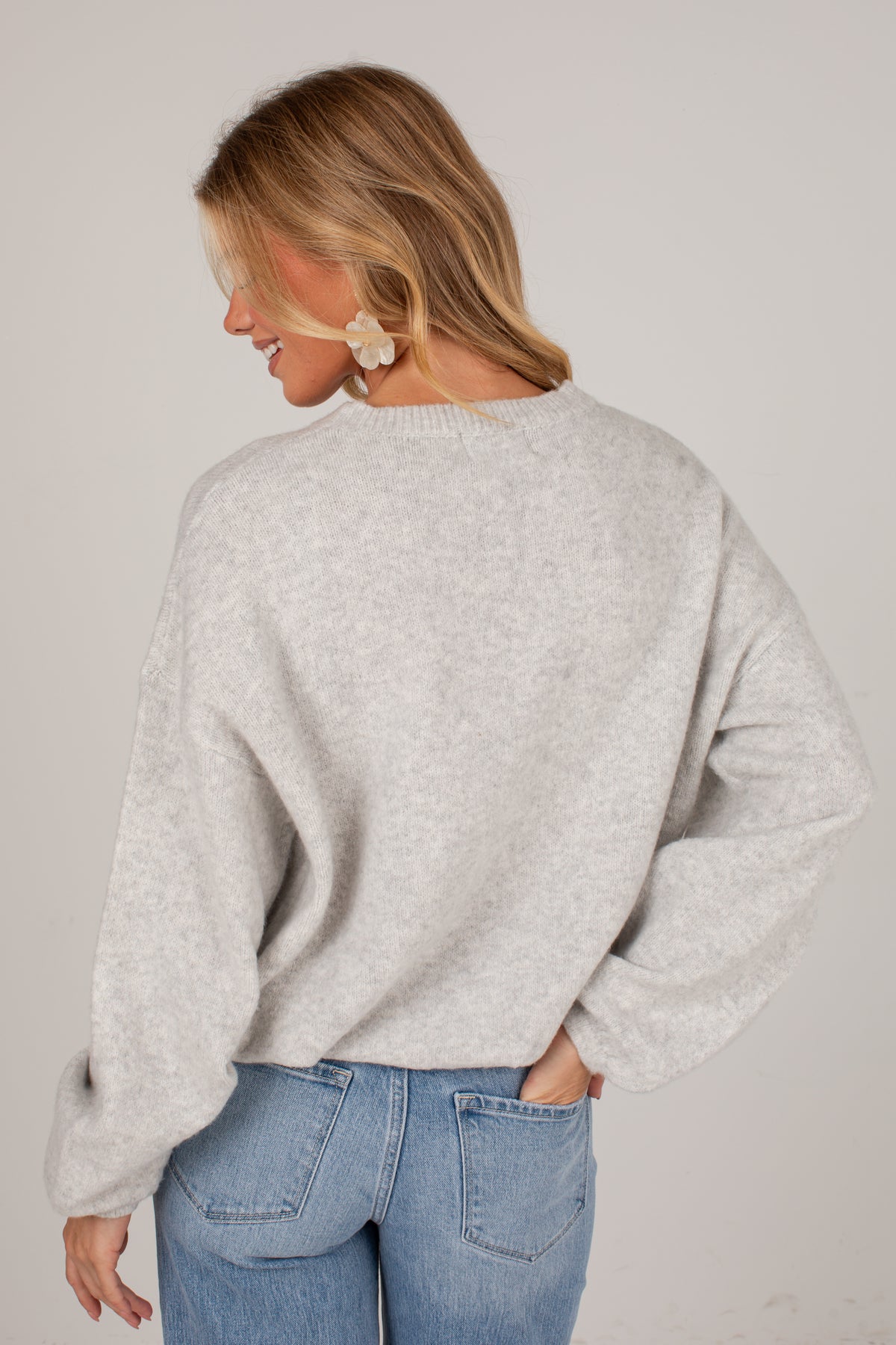 A woman shown from the back wearing a light heather gray knit sweater and blue jeans against a plain light gray studio background.