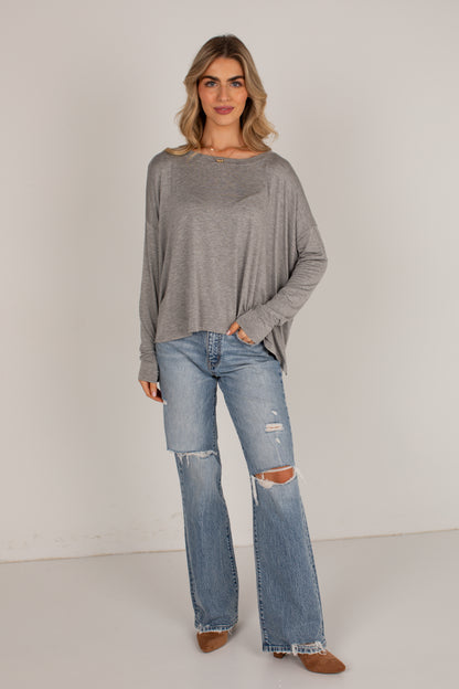 A woman modeling a loose-fit, long-sleeve gray knit top paired with light-wash distressed flare jeans and brown boots, standing against a plain light gray studio background.