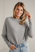 A woman wearing a light gray long-sleeve knit top and light-wash blue jeans, standing against a white paneled wall.