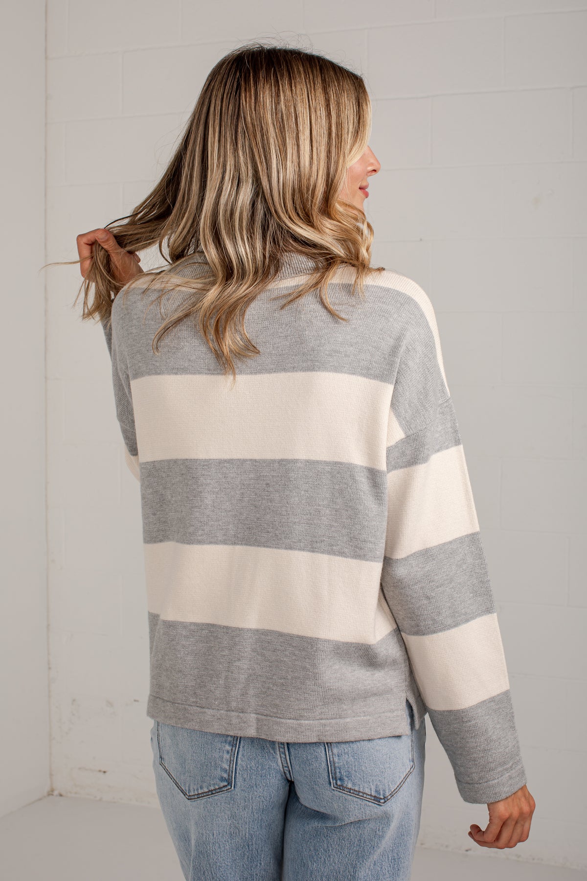A woman shown from the back wearing a gray and cream striped long-sleeve sweater with light blue jeans, standing against a white paneled wall.