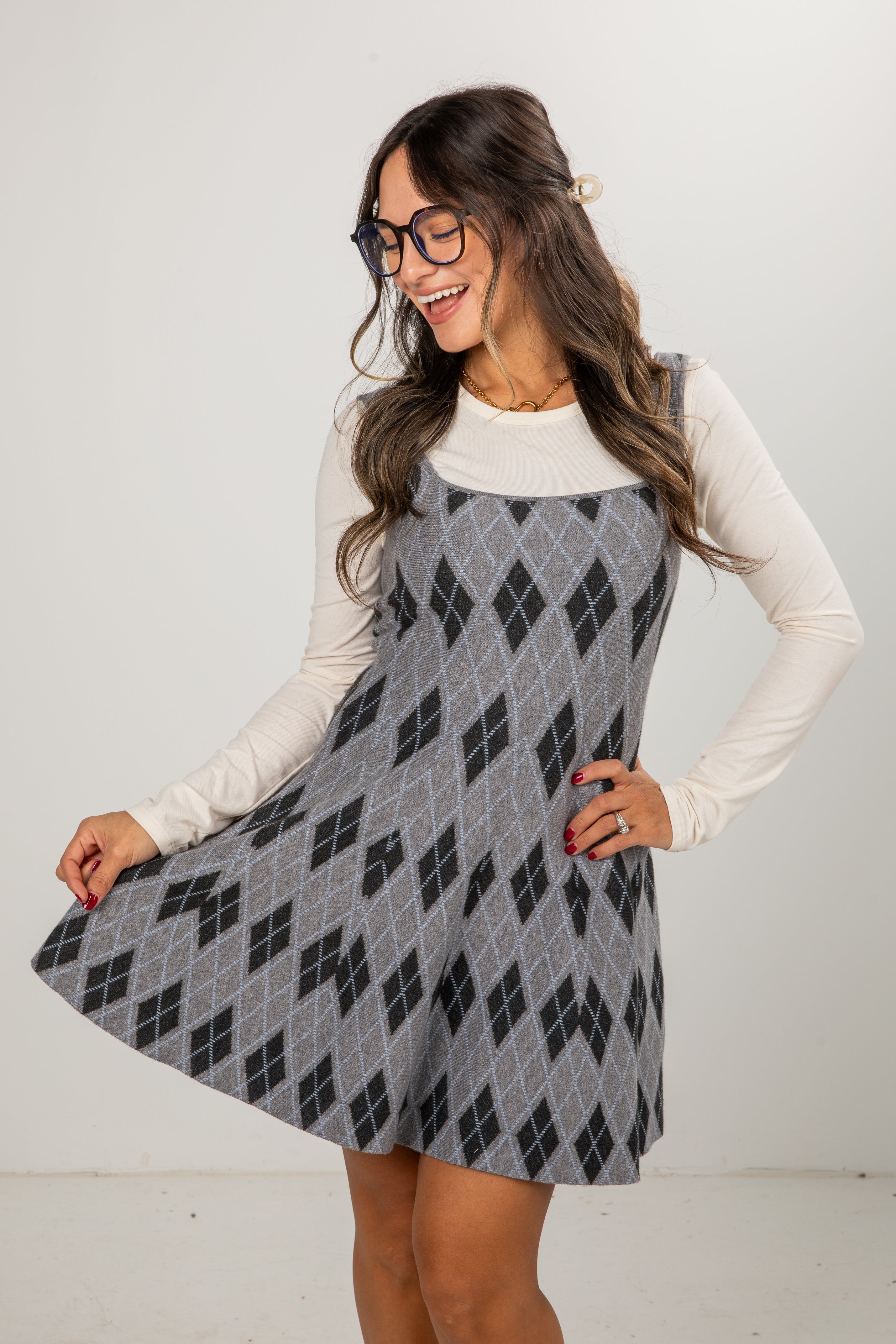 Woman wearing a gray patterned dress with a white long-sleeve shirt underneath on a plain background