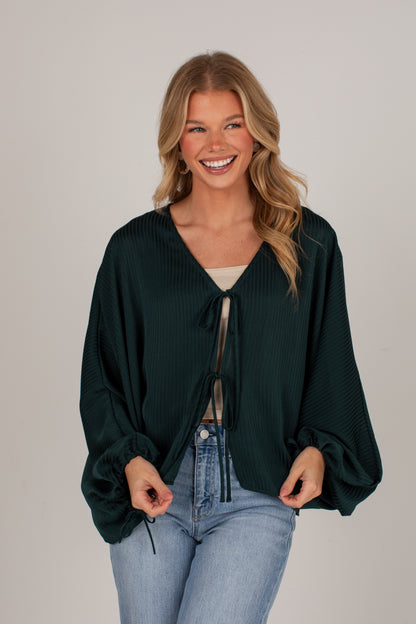 A smiling blonde woman wearing a dark forest green textured long-sleeve wrap top with a tie-front detail, styled with light-wash blue jeans against a plain light gray studio background.