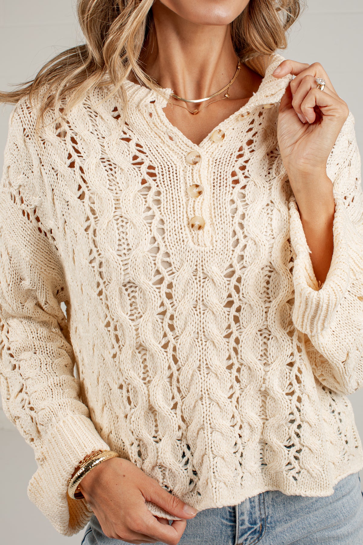 A woman wearing a cream-colored, open-knit long-sleeve sweater with a buttoned V-neck, paired with blue jeans and gold jewelry, against a neutral gray background.