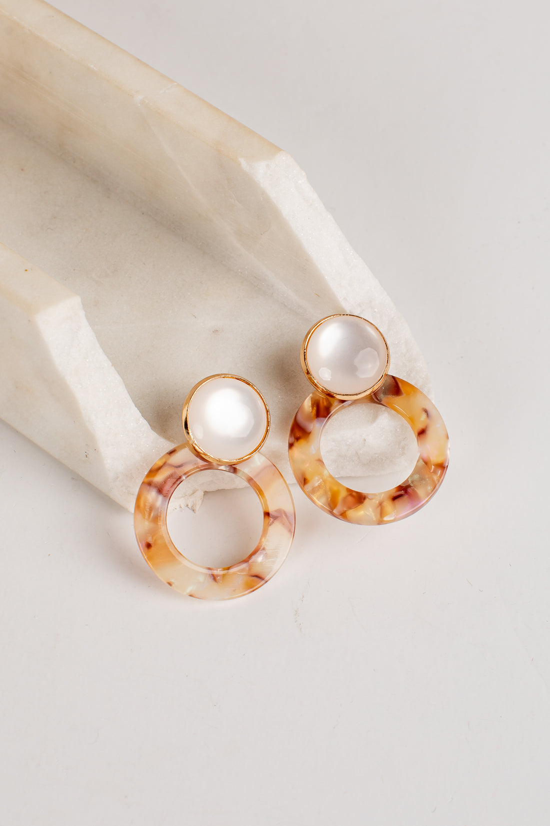 A pair of drop earrings with round white faux pearls at the top and large tortoiseshell acetate rings below, displayed on a cream-colored stone block against a light gray background.