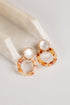 A pair of drop earrings with round white faux pearls at the top and large tortoiseshell acetate rings below, displayed on a cream-colored stone block against a light gray background.