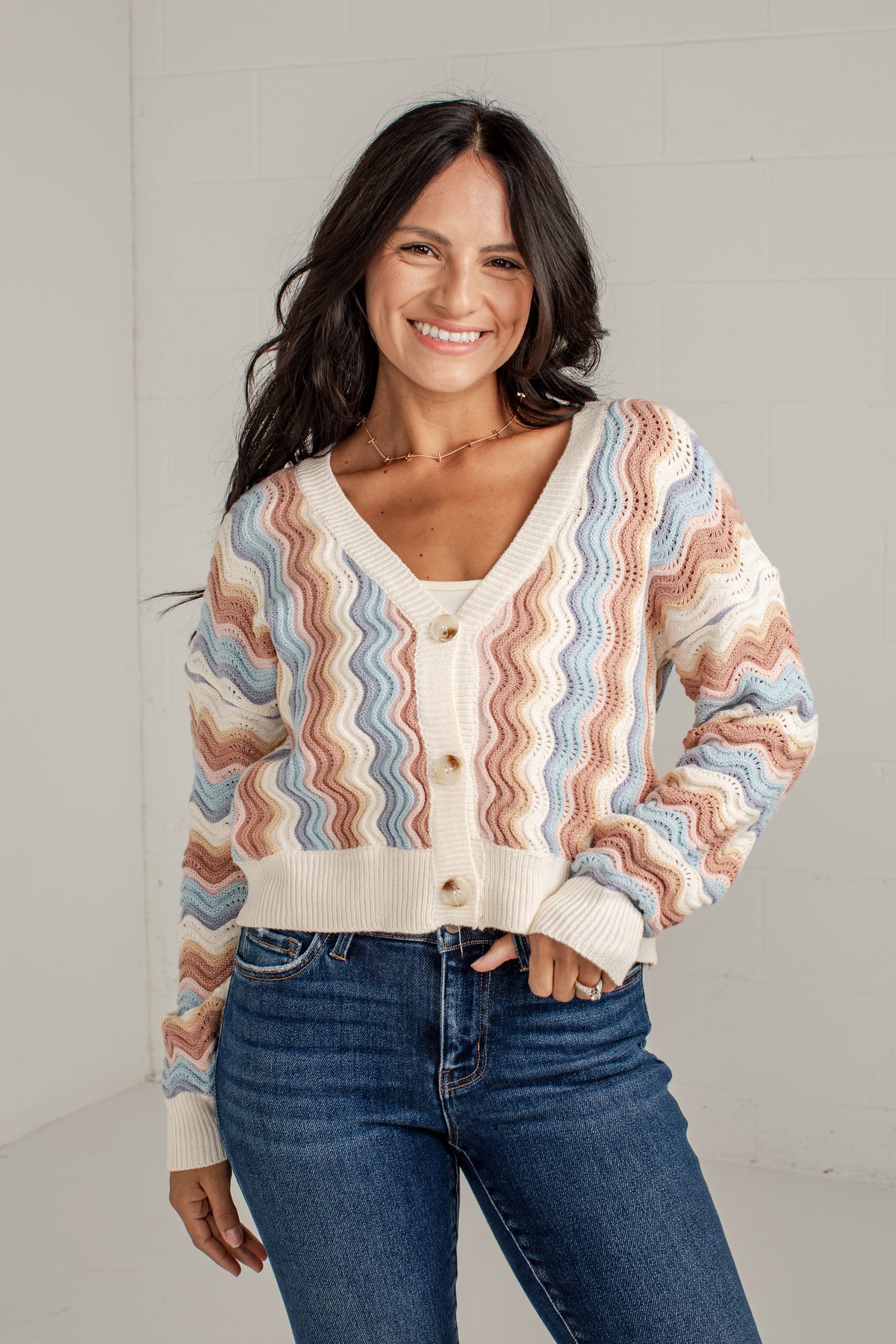 A woman wearing a cream-colored knit cardigan with a wavy, zigzag pattern in dusty pink, light blue, and terracotta, paired with blue jeans, standing against a white brick wall.