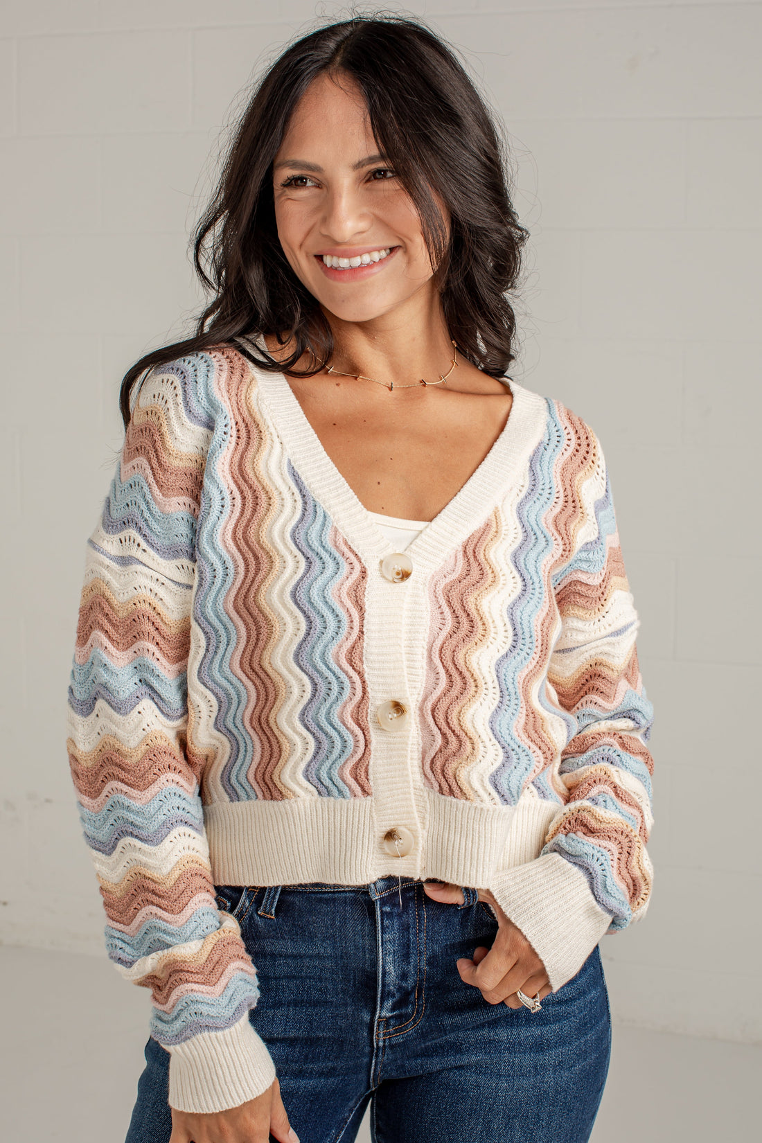A woman wearing a cream knit cardigan with wavy pastel stripes in blue, pink, and tan, paired with dark blue jeans, standing against a light gray wall.