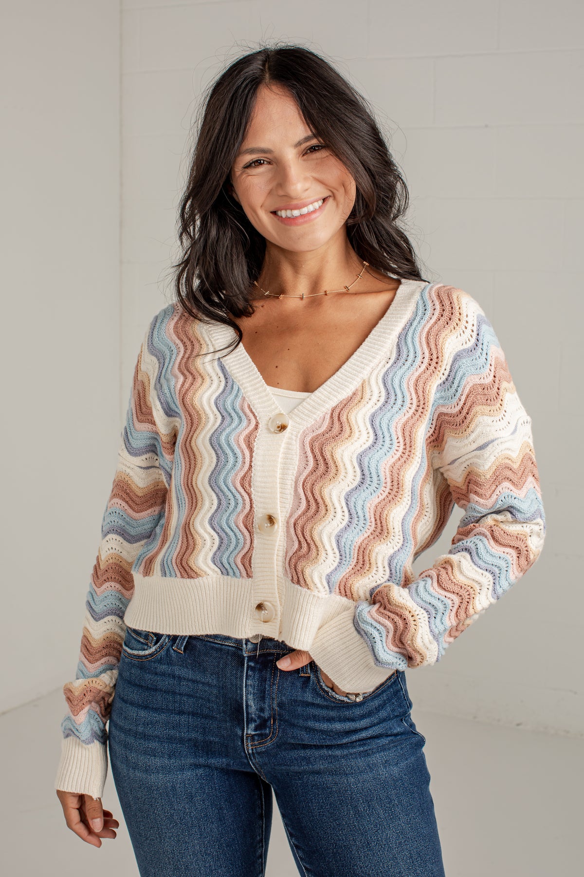 A smiling woman wearing a cream knit cardigan with wavy pastel stripes in blue, pink, and tan, paired with blue jeans, standing against a light gray wall.