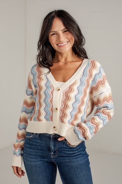 A smiling woman wearing a cream knit cardigan with wavy pastel stripes in blue, pink, and tan, paired with blue jeans, standing against a light gray wall.