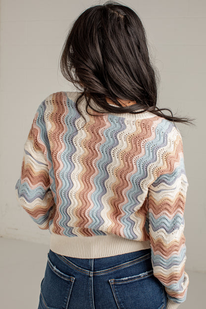 A person shown from the back wearing a cropped knit sweater with a wavy multicolor stripe pattern in beige, blue, pink, and brown, paired with blue jeans against a plain light wall.