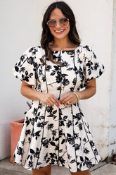 Woman wearing a black floral dress standing against a white wall.