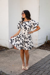 Woman wearing a black and white floral dress standing against a white wall.