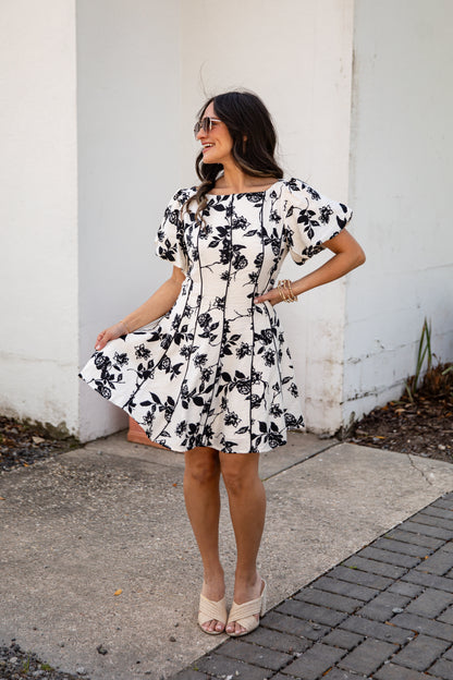 Woman wearing a black and white floral dress standing against a white wall.