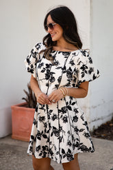 Woman wearing a black and white floral dress standing against a white wall.