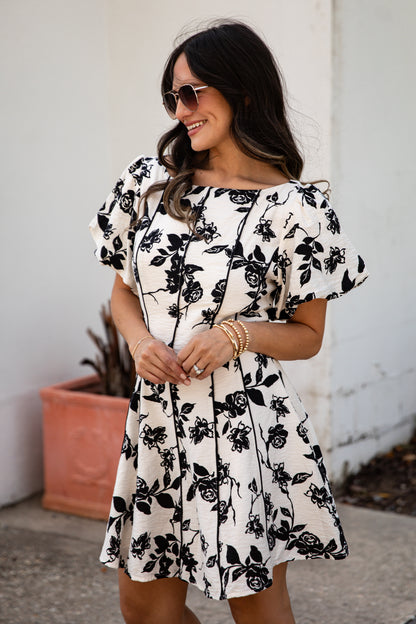 Woman wearing a black and white floral dress standing against a white wall.