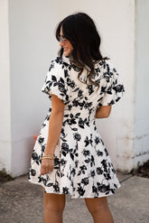 Woman wearing a black and white floral dress against a white wall.