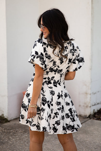 Woman wearing a black and white floral dress against a white wall.