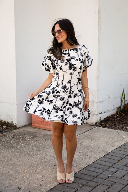 Woman wearing a black and white floral dress standing against a white wall.