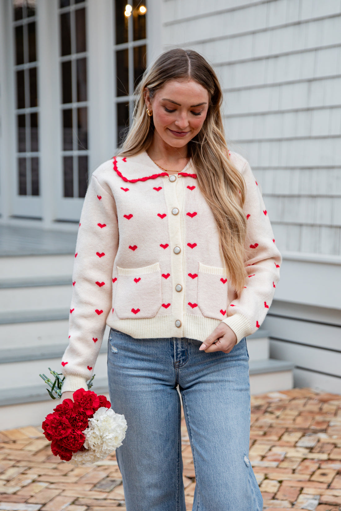 Woman wearing a cream cardigan with red heart patterns, holding flowers, standing on a brick path.