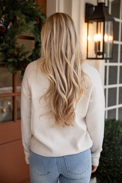 Person wearing a beige sweater and blue jeans with long blonde hair, standing in front of a house entrance.