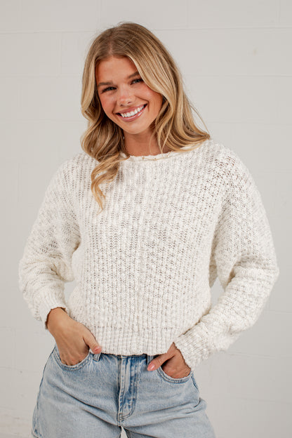 A smiling blonde woman wearing a chunky knit cream-colored sweater and light-wash blue jeans, standing against a light gray wall.