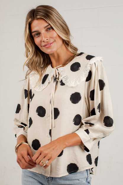 A woman wearing a cream-colored long-sleeve blouse with black polka dots and a lace-trimmed ruffled collar, paired with light blue jeans, standing against a white brick wall.