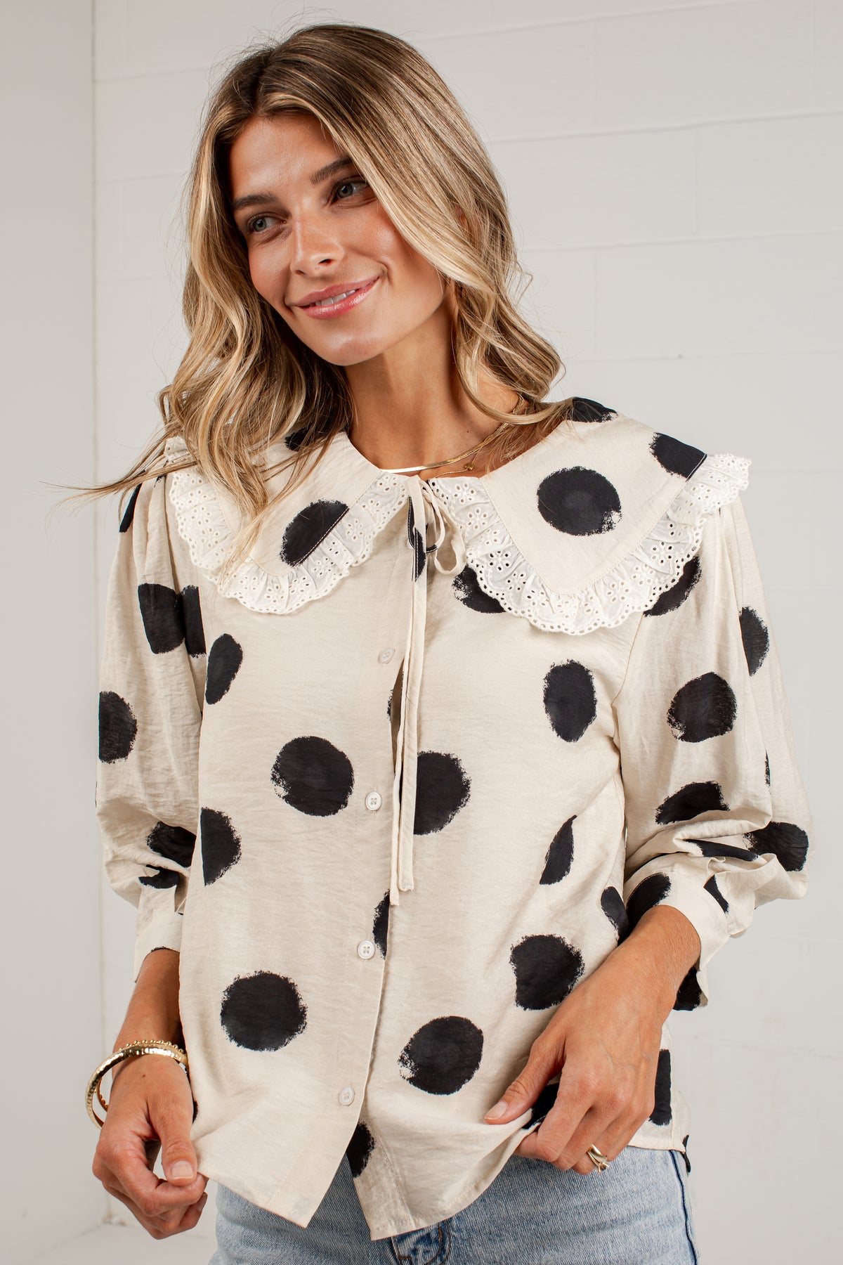 A woman wearing a cream-colored long-sleeve blouse with black polka dots, a lace-trimmed Peter Pan collar, and a front tie closure, paired with light blue jeans.