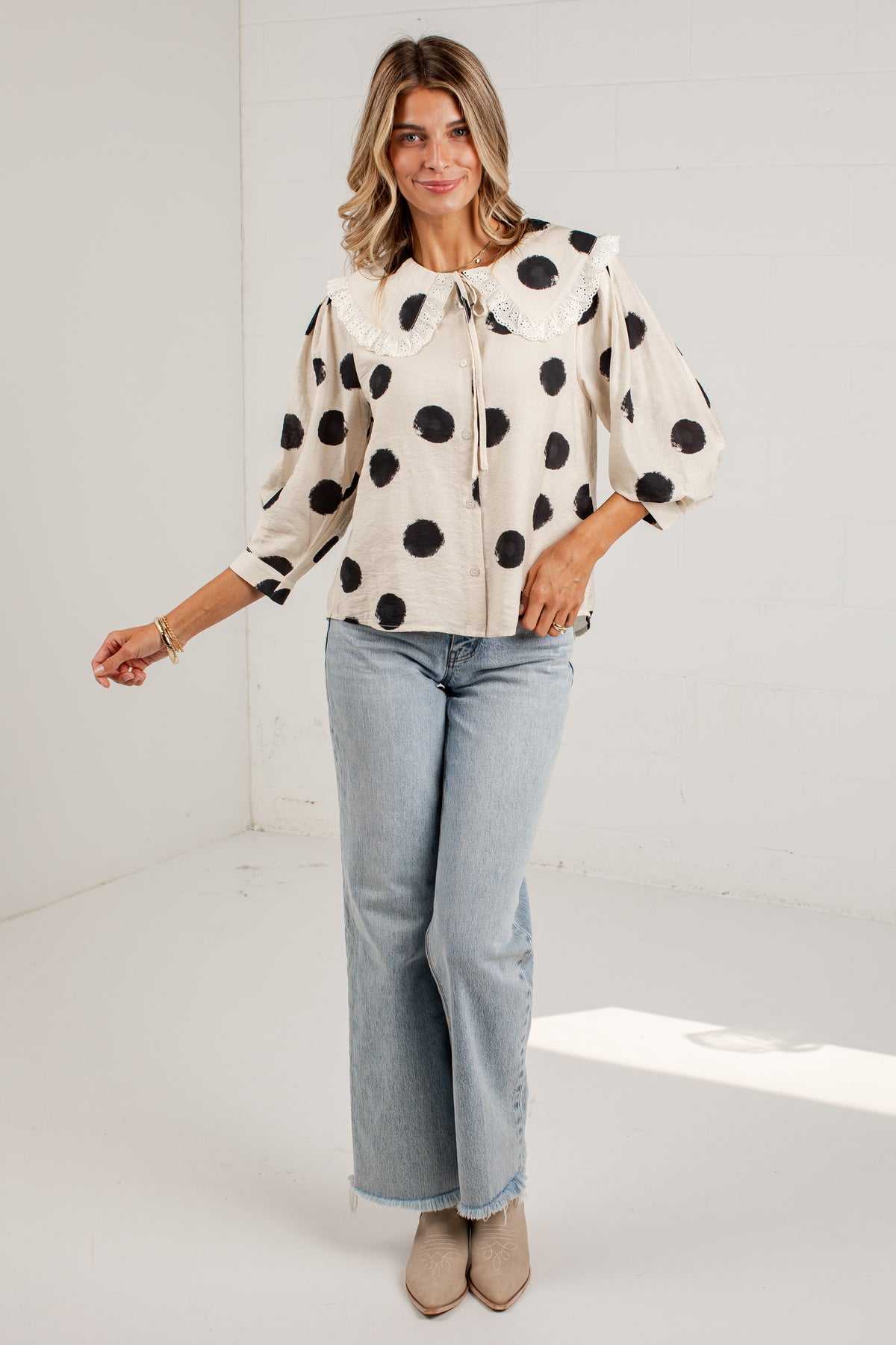 A woman modeling a cream-colored blouse with black polka dots, a large Peter Pan collar, and puff sleeves, paired with light-wash blue jeans and beige ankle boots, in a bright white studio setting.
