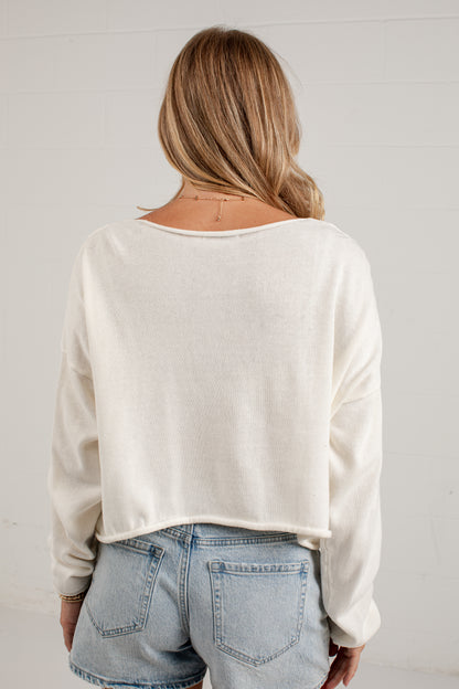 A woman viewed from behind wearing a cream-colored, long-sleeve knit crop top and light-wash denim shorts against a white paneled wall.