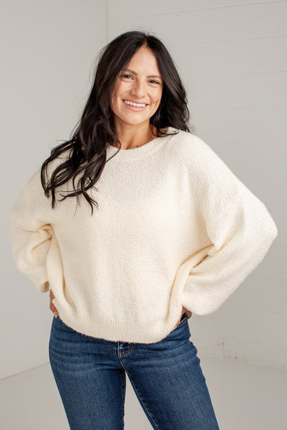 A smiling woman with long dark hair wears a cream-colored, textured bubble-sleeve knit sweater and blue jeans, standing against a light gray wall.