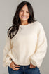 A smiling woman with long dark hair wearing a cream-colored, textured bubble-sleeve knit sweater and blue jeans, standing against a light gray wall.