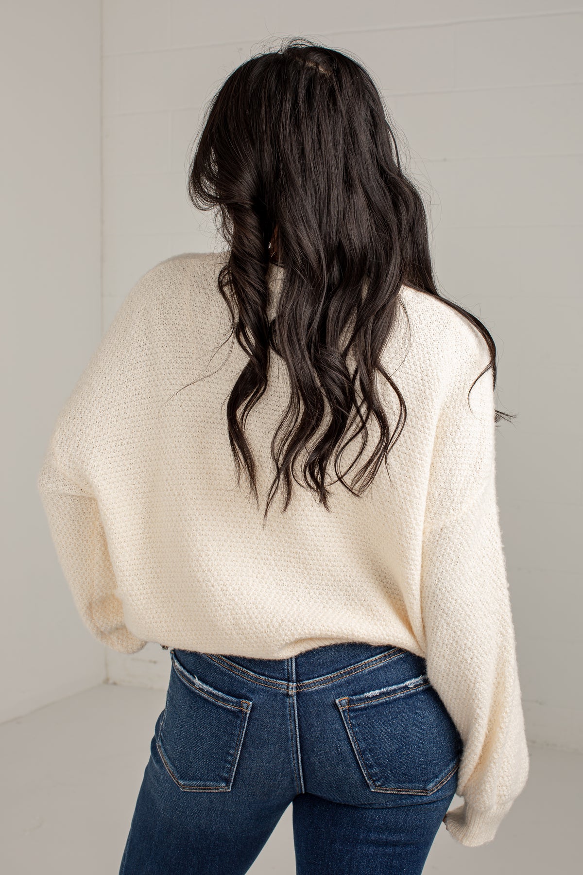A woman viewed from behind wearing a cream-colored, textured knit sweater and blue jeans against a white paneled wall.