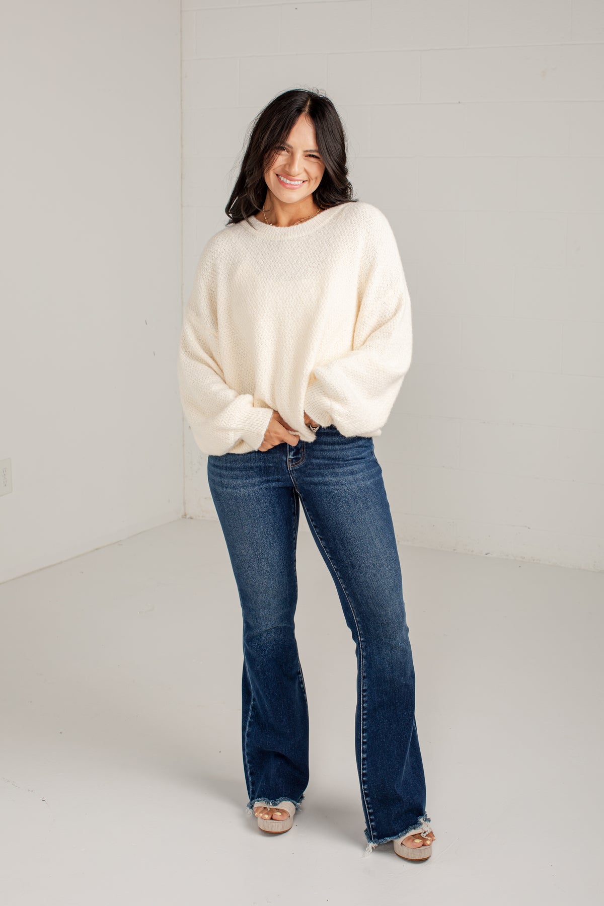 A woman modeling a cream-colored, oversized knit sweater and dark blue flared jeans against a plain white studio background.