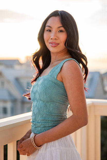 Woman in a teal top and white skirt standing on a balcony with a blurred cityscape background