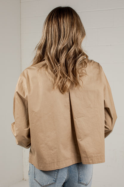 A woman viewed from behind wearing a light tan utility-style jacket and light blue jeans, standing against a white brick wall.
