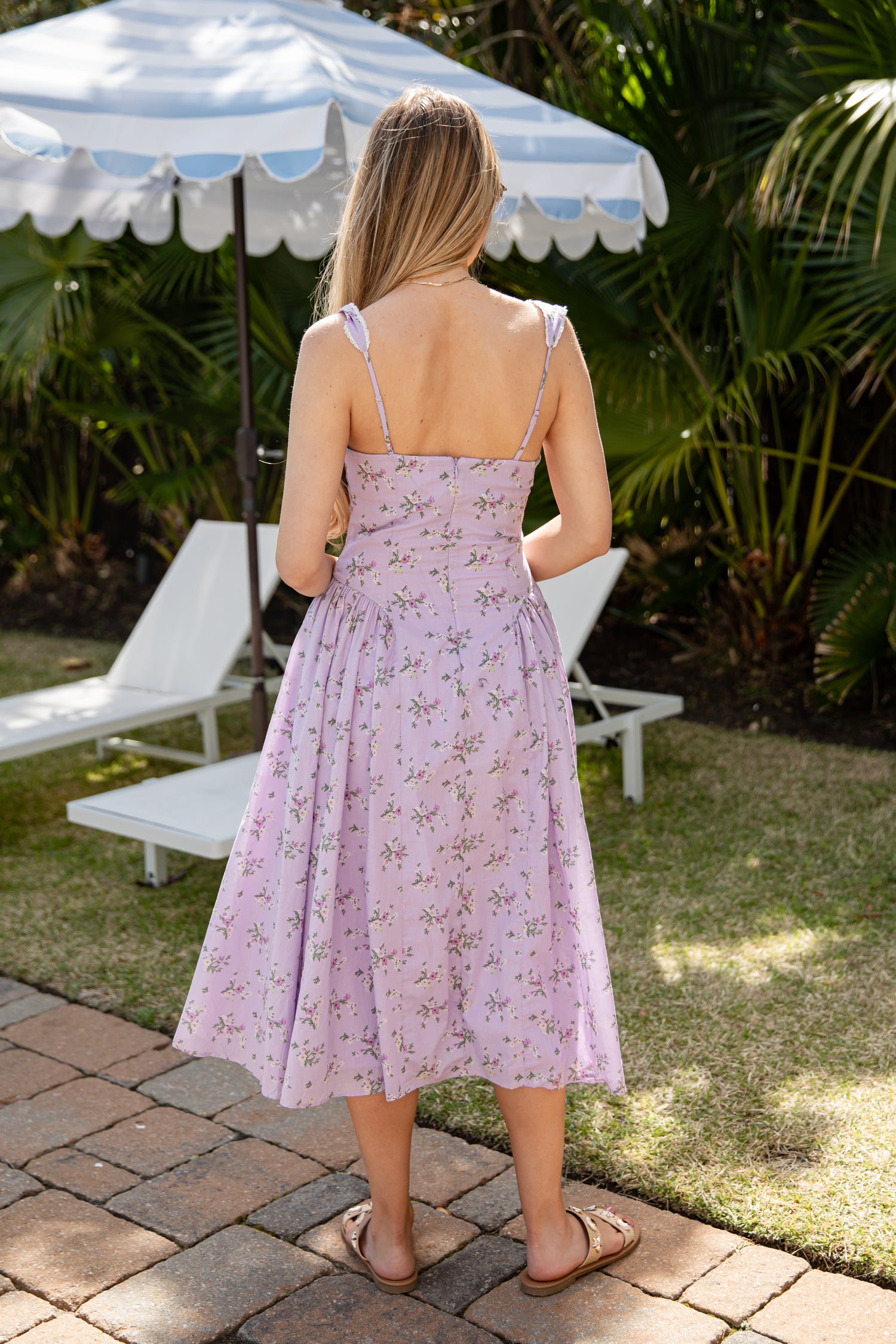 Woman in a pink floral dress standing outdoors under a striped umbrella with lounge chairs and greenery.