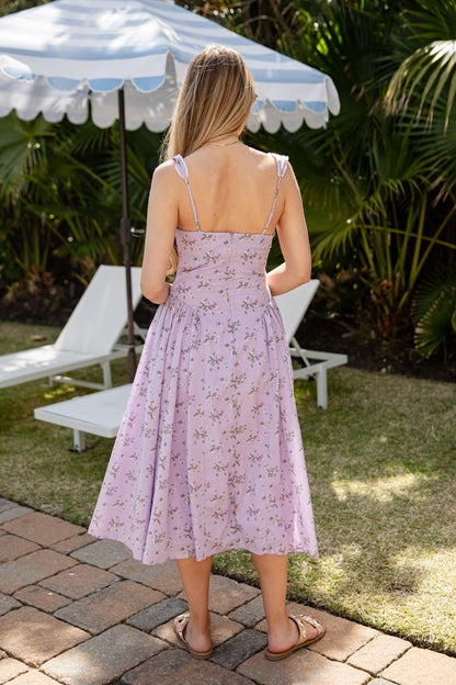 Woman in a pink floral dress standing outdoors under a striped umbrella with lounge chairs and greenery.