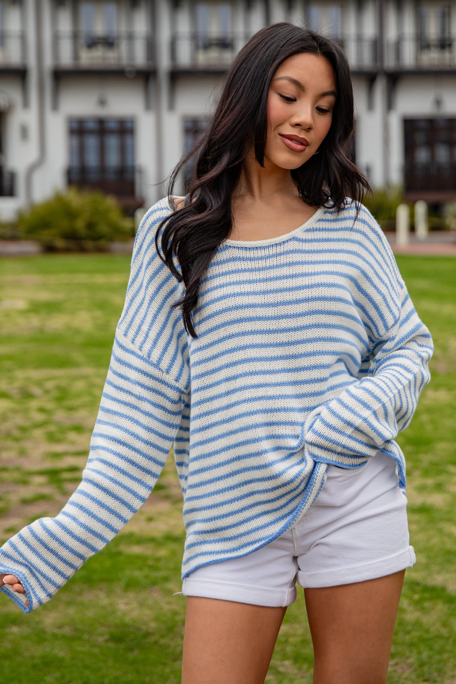 Woman wearing a blue and white striped sweater and white shorts standing in a grassy area with a building in the background.