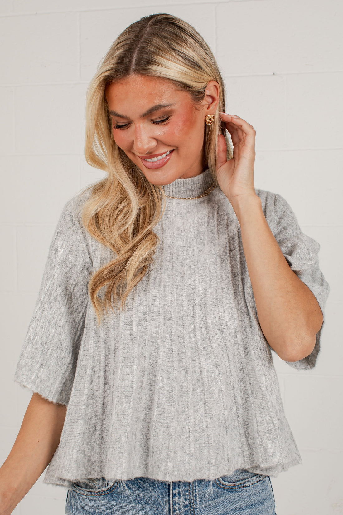 A woman wearing a light gray heathered short-sleeve mock neck top with a textured crinkle fabric, paired with blue jeans, standing against a white brick wall.
