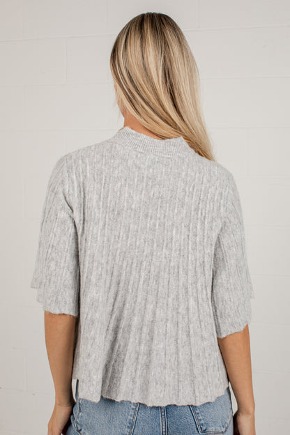 A woman viewed from behind wearing a light gray ribbed knit short-sleeve top and blue jeans against a white brick wall.