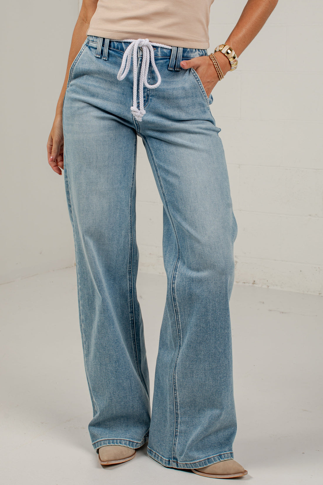 A model wearing light-wash blue wide-leg jeans with a white drawstring waist, paired with a beige top and beige shoes, standing against a white studio background.